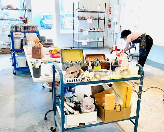 an artist in a large room with art supplies on a cart and a bag of clay on the table