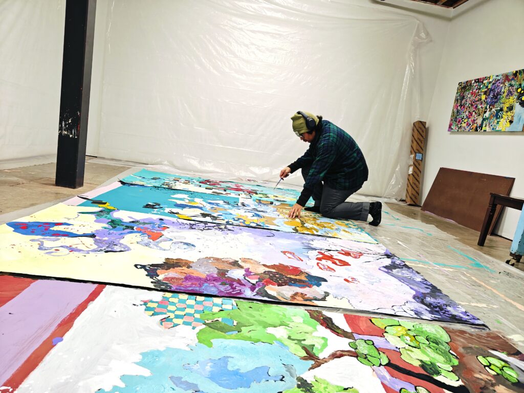 an artist kneels on the floor in front of vibrant works in progress with a paintbrush in their hand. he wears a beanie, headphones, plaid shirt, grey jeans, and socks.