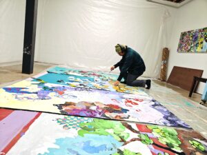 an artist kneels on the floor in front of vibrant works in progress with a paintbrush in their hand. he wears a beanie, headphones, plaid shirt, grey jeans, and socks.