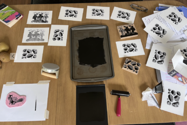 A wooden table covered with prints of black leaf patterns and ink stamps, arranged around a central ink tray and stamping tools.