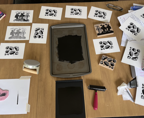 A wooden table covered with prints of black leaf patterns and ink stamps, arranged around a central ink tray and stamping tools.