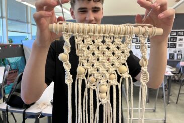 Person demonstrates macrame weaving, holding a wooden dowel with knotted cream cords and wooden beads in a classroom.