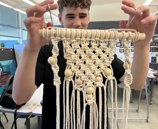 Person demonstrates macrame weaving, holding a wooden dowel with knotted cream cords and wooden beads in a classroom.