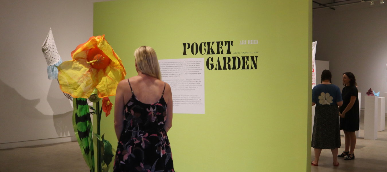 Woman in a floral dress stands before a lime-green wall labeled 'Pocket Garden' with a large orange-yellow flower sculpture to her left in a gallery.