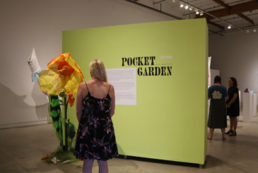 Woman in a floral dress stands before a lime-green wall labeled 'Pocket Garden' with a large orange-yellow flower sculpture to her left in a gallery.