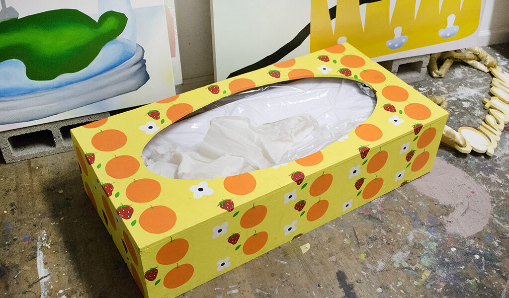 Yellow tissue box with orange fruit pattern and exposed tissues on a paint-splattered floor in an art studio.