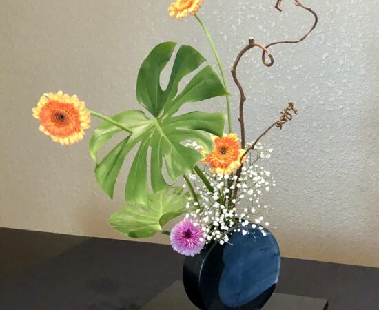 Colorful flower arrangement with orange gerberas, white baby’s breath, green leaves, and curly branches in a blue vase on a dark table surface.