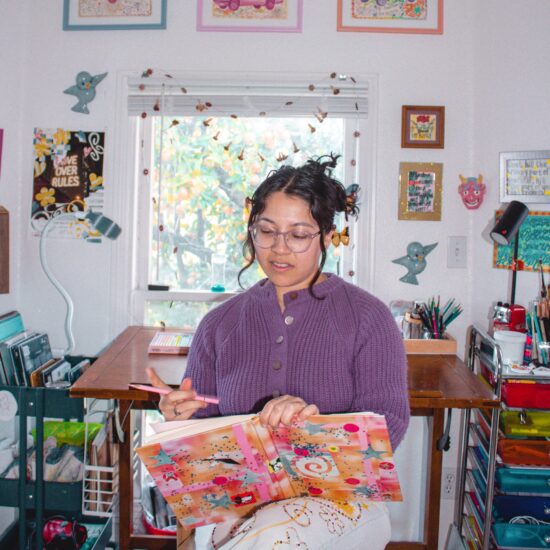 Woman in a purple sweater sits at a craft table, decorating a colorful gift bag with stars and swirls in a bright room.
