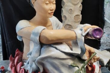 Seated ceramic sculpture of a woman with a pale blue robe, holding a purple cup, surrounded by red lotus petals at the base.