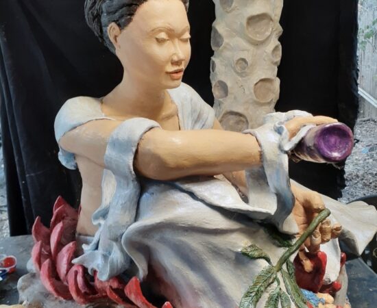 Seated ceramic sculpture of a woman with a pale blue robe, holding a purple cup, surrounded by red lotus petals at the base.