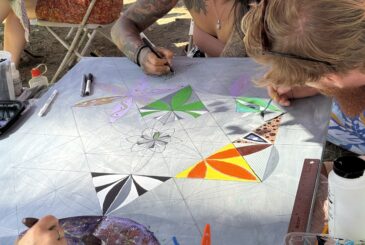 Two artists sketch geometric patterns on a large canvas at an outdoor art table under an orange canopy.