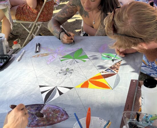 Two artists sketch geometric patterns on a large canvas at an outdoor art table under an orange canopy.
