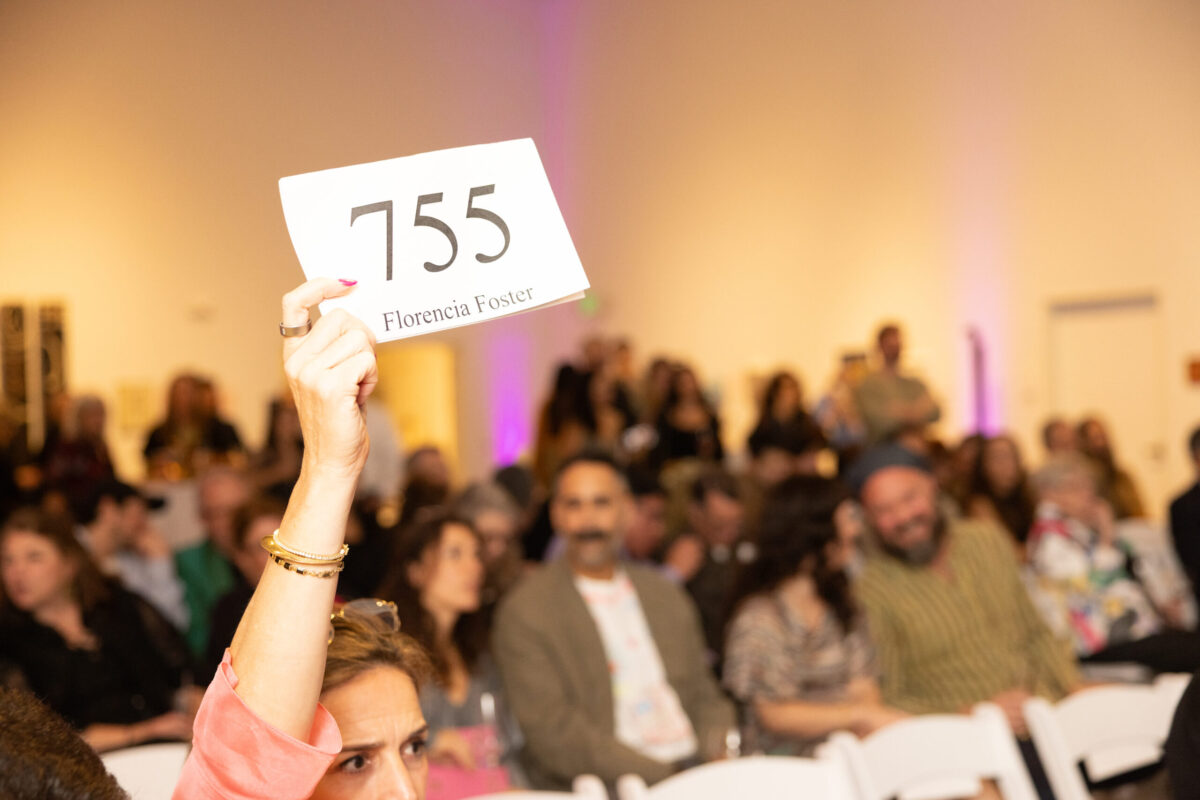 Raised white sign displaying '155' and 'Florencia Foster' held above a crowded indoor event.