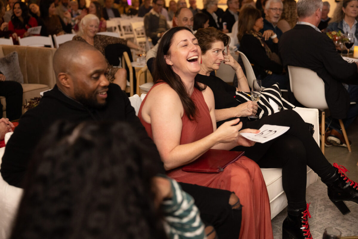 Laughing woman in a red dress seated on a white sofa at a social event; others smile and chat in the background with wine glasses nearby.
