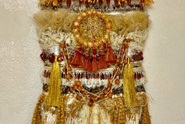 Decorative textile wall hanging with white, cream, and gold strips, beads, and long gold tassels forming a layered sculpture on a neutral wall.