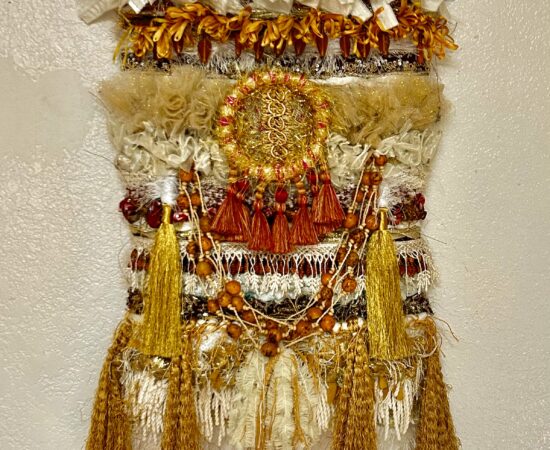 Decorative textile wall hanging with white, cream, and gold strips, beads, and long gold tassels forming a layered sculpture on a neutral wall.