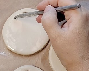 Close-up of a hand using a metal modeling tool to poke a hole in a white clay disk on a work surface