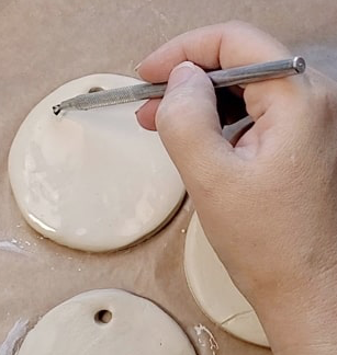 Close-up of a hand using a metal modeling tool to poke a hole in a white clay disk on a work surface
