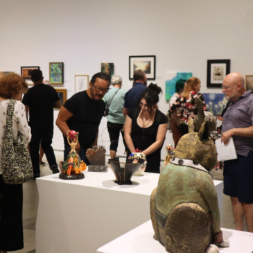 Visitors in an art gallery examine ceramic sculptures on white pedestals, with a large green-brown animal statue with long ears in the foreground.