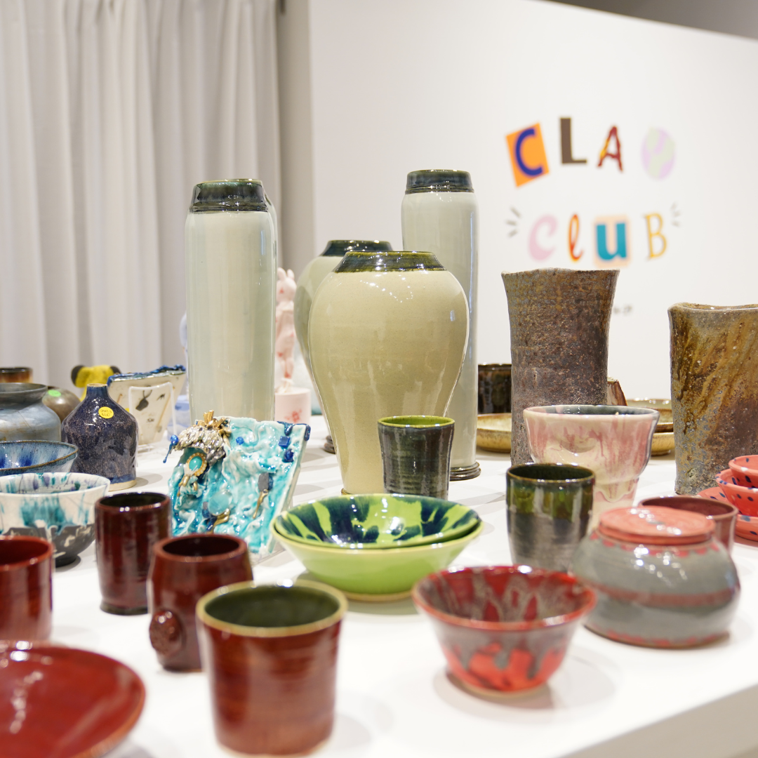 1 - Verge Center for the Arts Assortment of glazed ceramic pottery—vases, bowls, and cups—on a white display table at a clay club exhibit, with a colorful sign in the background.